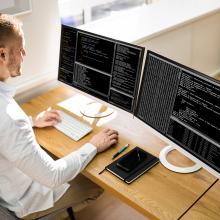 Una mujer sentada frente a dos pantallas de ordenador, gestionando la seguridad de la informaci&oacute;n.