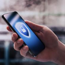 Unas manos sostienen un tel&eacute;fono m&oacute;vil, en cuya pantalla aparece el icono de un escudo, que represnta las buenas pr&aacute;cticas frente al phishing.