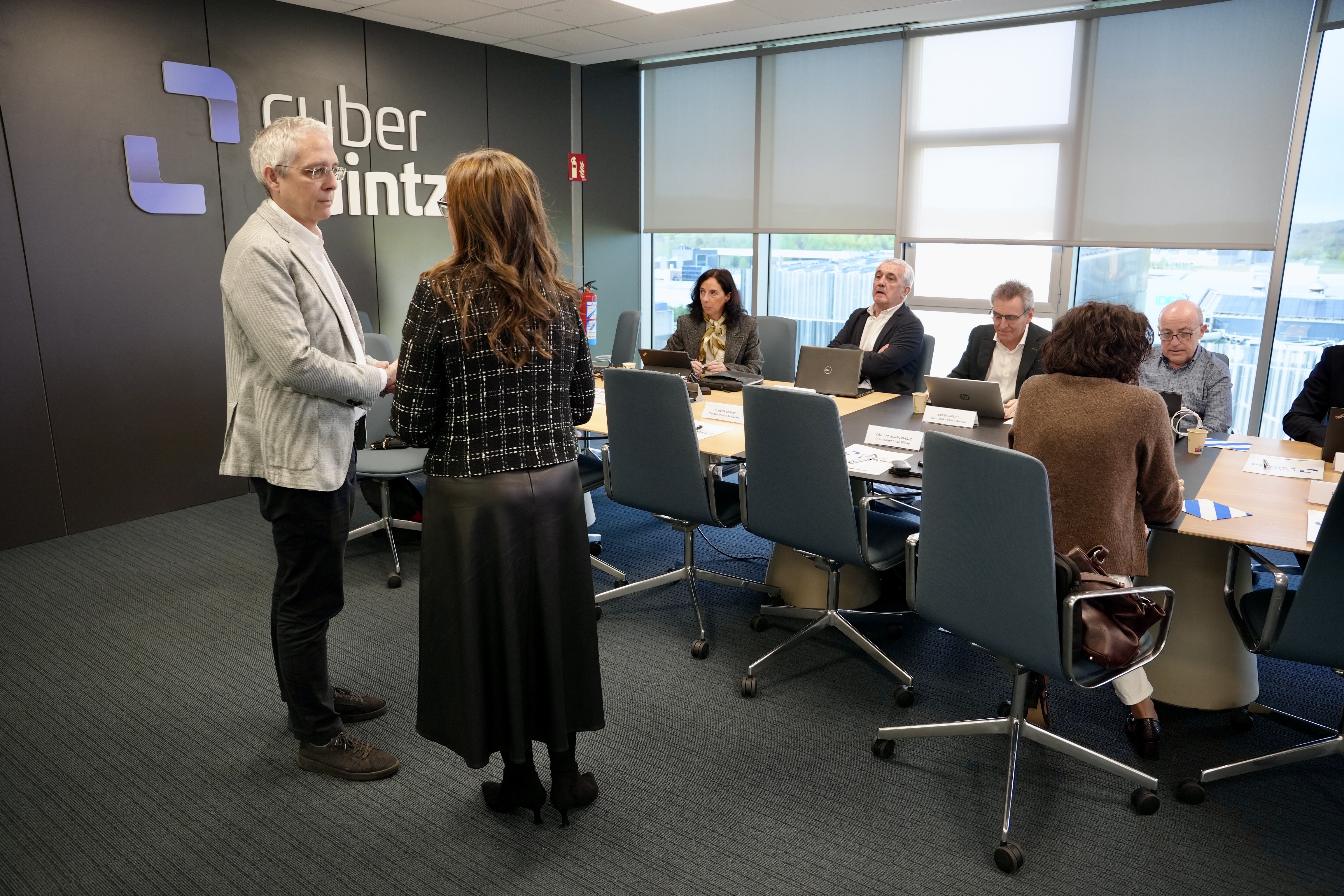 Foto de un momento de la reuni�n del Consejo de Administraci�n de Cyberzaintza, en el que dos participantes se ven de pie y otros sentados en la mesa de reuniones de la Agencia Vasca de Ciberseguridad.